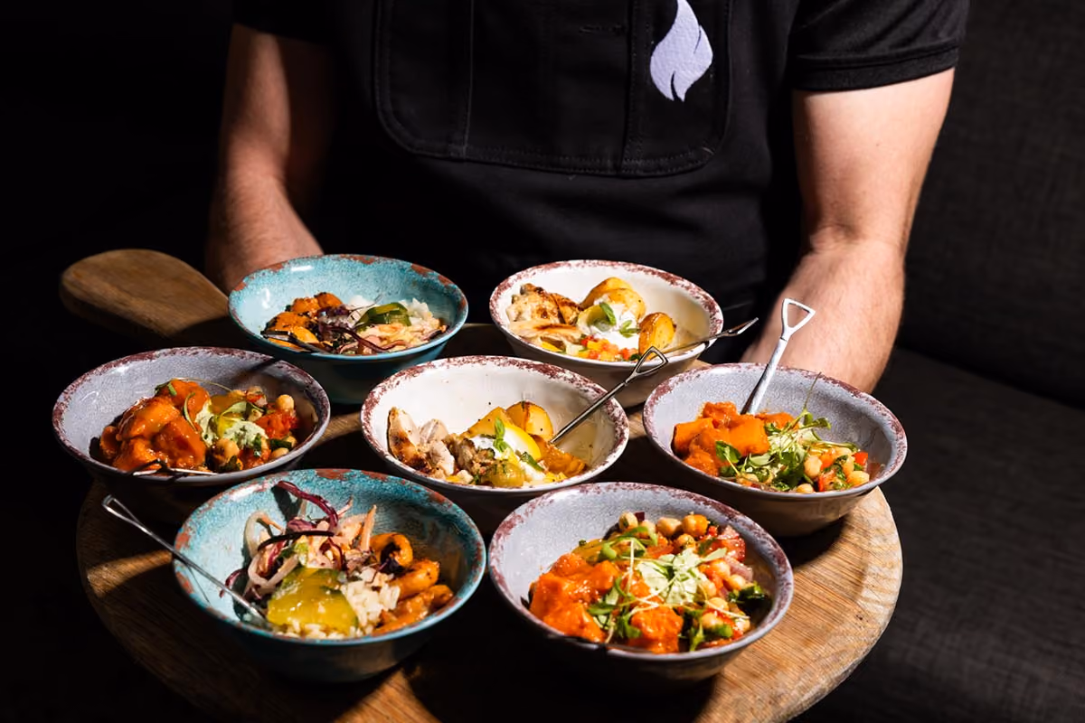 Nyama bowls of assorted grilled chicken, roasted potatoes, chickpea salad, and vegetables on a round wooden tray.