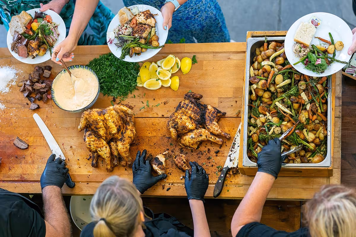 Nyama staff serving grilled whole chickens, roasted potatoes, and vegetables onto plates at a food counter.