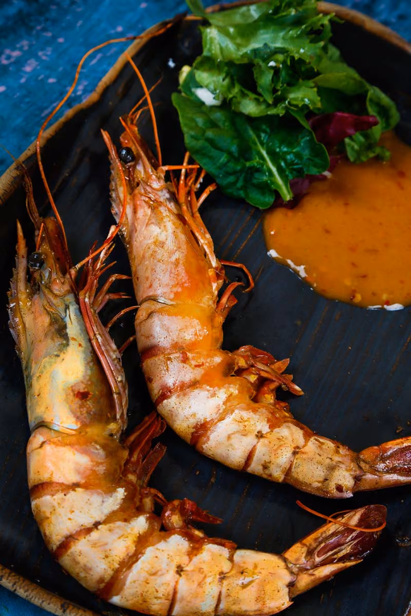 Two grilled shrimp with leafy greens and dipping sauce on a black plate.