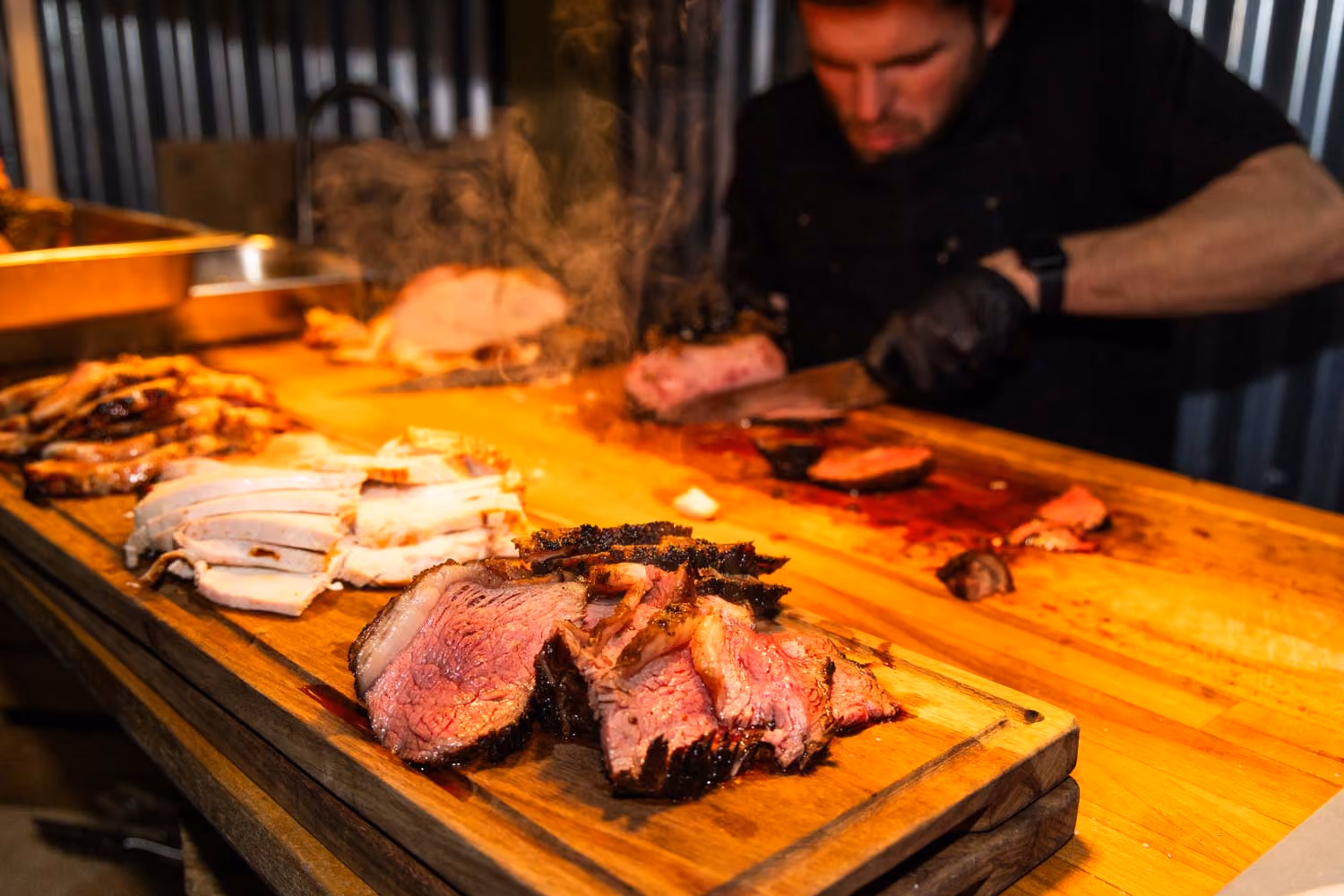 Cook slicing different smoked meats on a wooden board with steam rising in warm lighting.