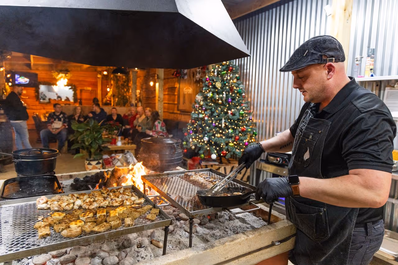 Man grilling meat near a Christmas tree with a group of people socializing in a cozy wooden room.