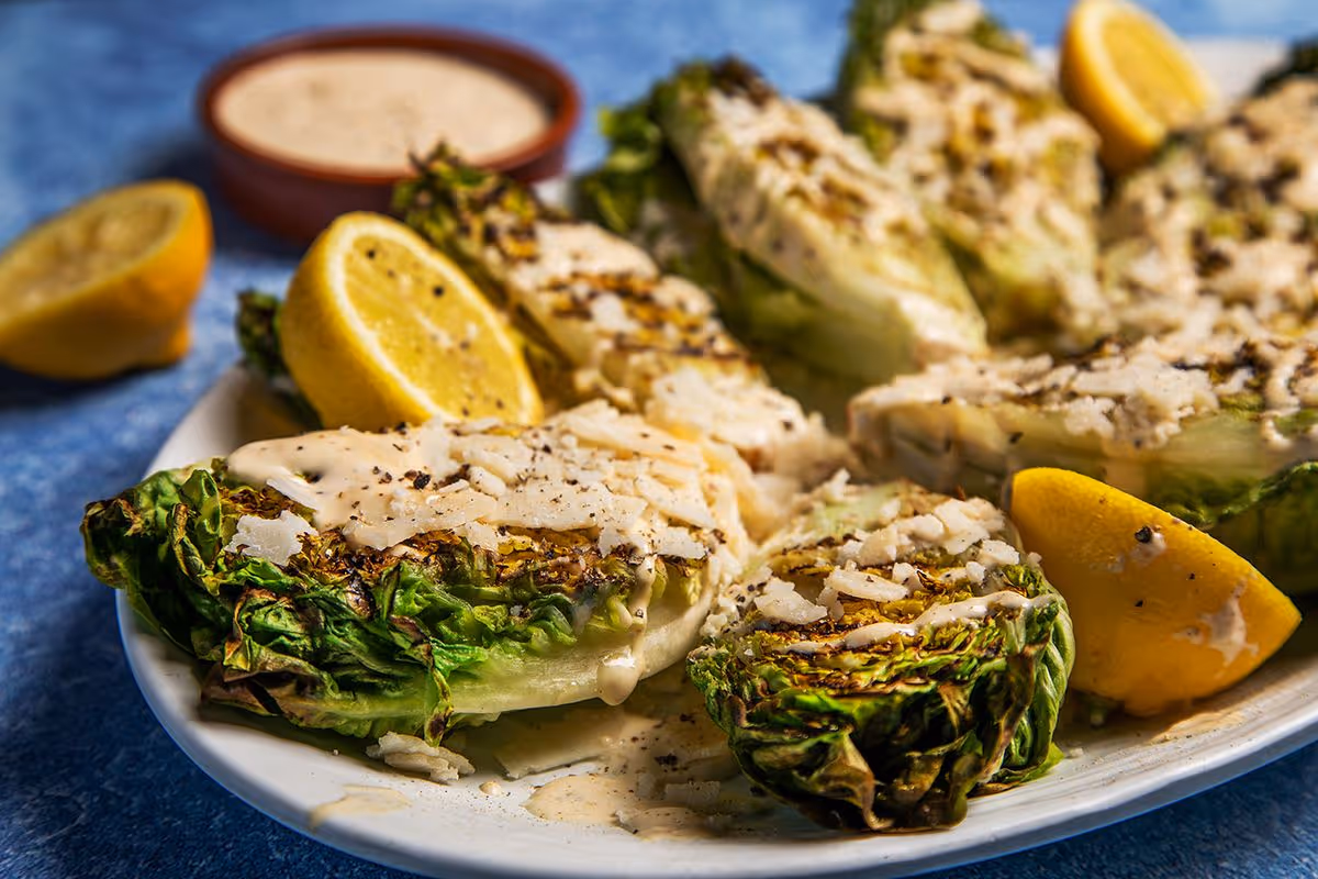 Grilled romaine lettuce wedges topped with creamy dressing, cheese, black pepper, and lemon wedges on a white plate.