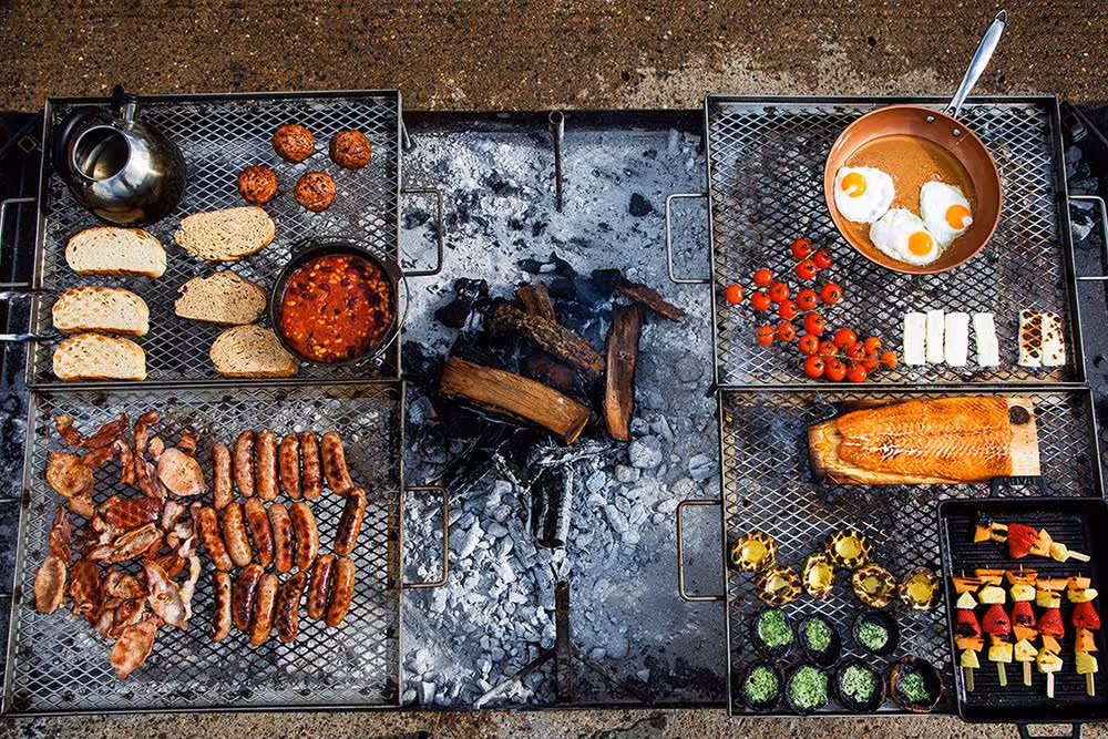 Outdoor campfire grill with sausages, bacon, eggs, bread, grilled fish, vegetables, and skewers cooking.