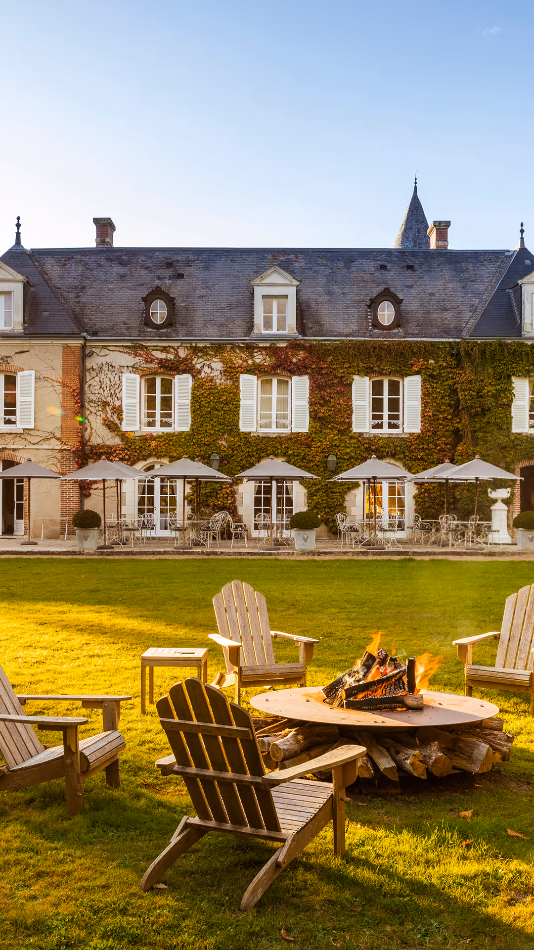 Wooden chairs around a campfire on lawn in front of a house with white shutters