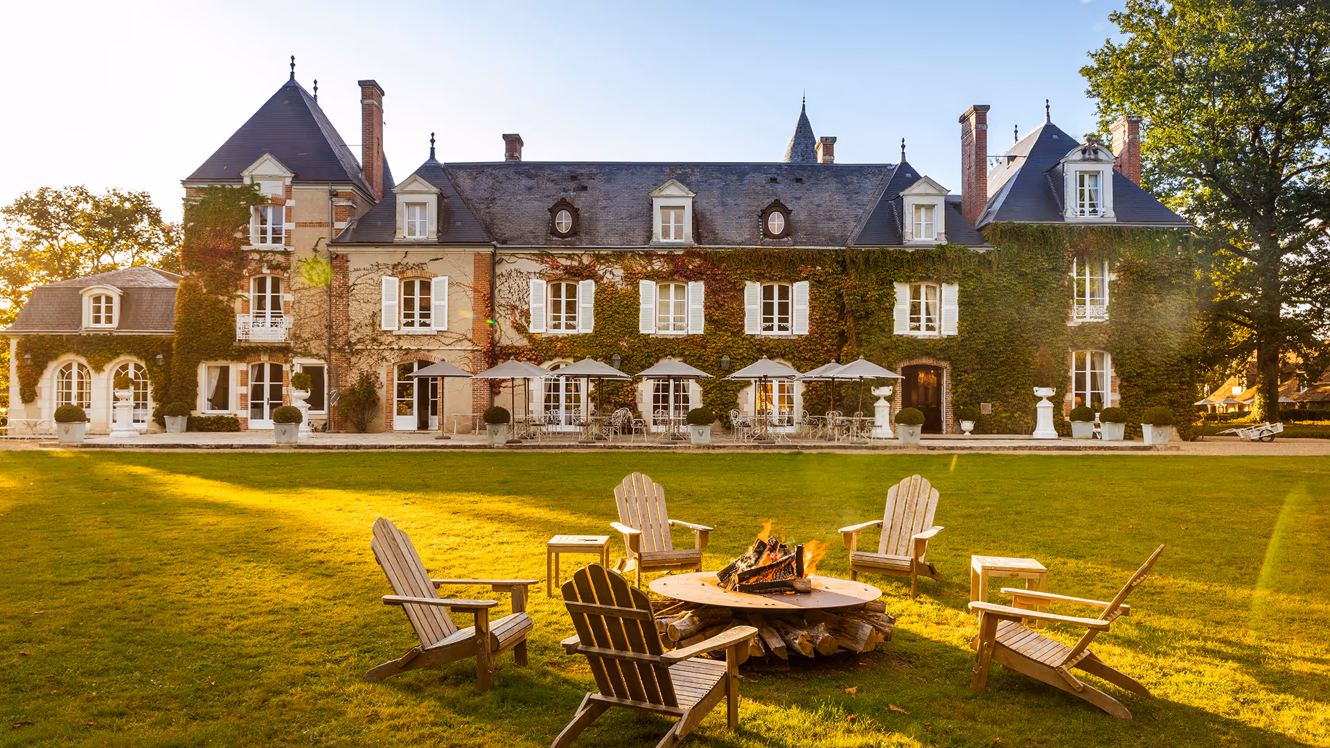 Large ivy-covered mansion with wooden chairs around a fire in a green lawn.