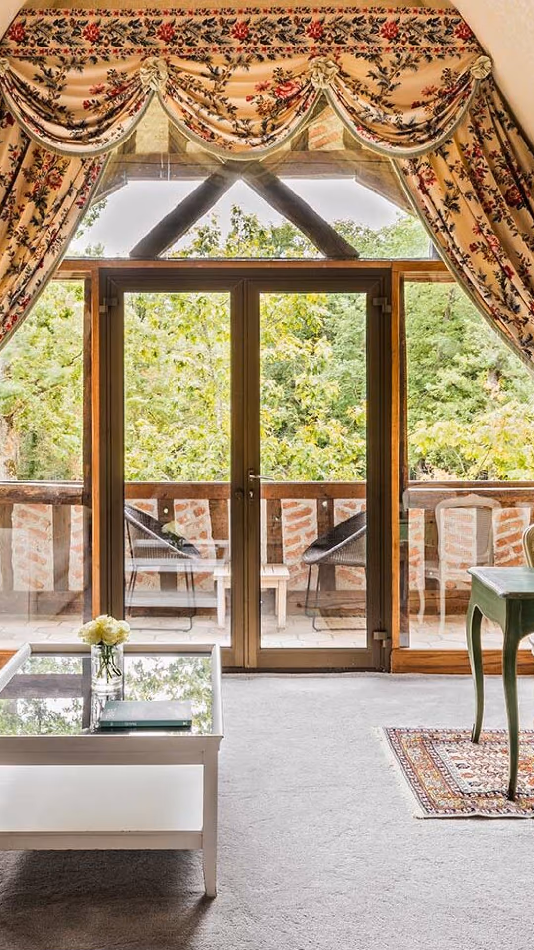 Glass door leading to a balcony with chairs, table, and views of green trees.