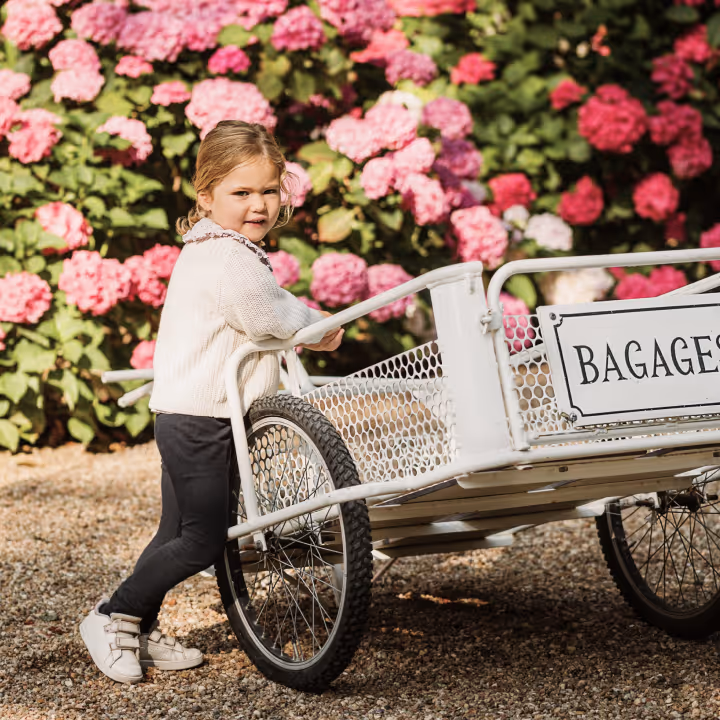 A little girl pushes a white cart marked BAGAGES in front of pink flowers.