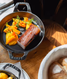 Dish with a piece of meat, orange purée and green toppings in a black pan.