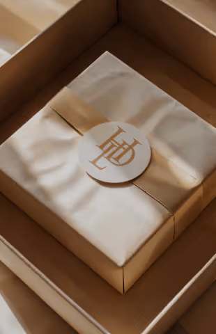 Beigefarbene Geschenkbox mit seidigem Papier und rundem Aufkleber mit goldenem LD-Logo.