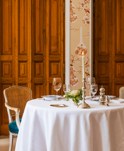 Round table set with white tablecloth, burning candle, glasses and a bouquet of flowers in an elegant room.