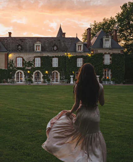 Woman in a flowing dress walking towards a large mansion covered in ivy at sunset.