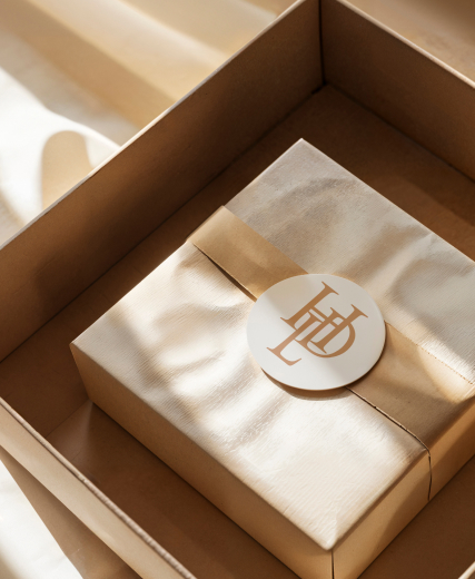 Beige gift box with a white and gold sticker with initials, in a cardboard box.