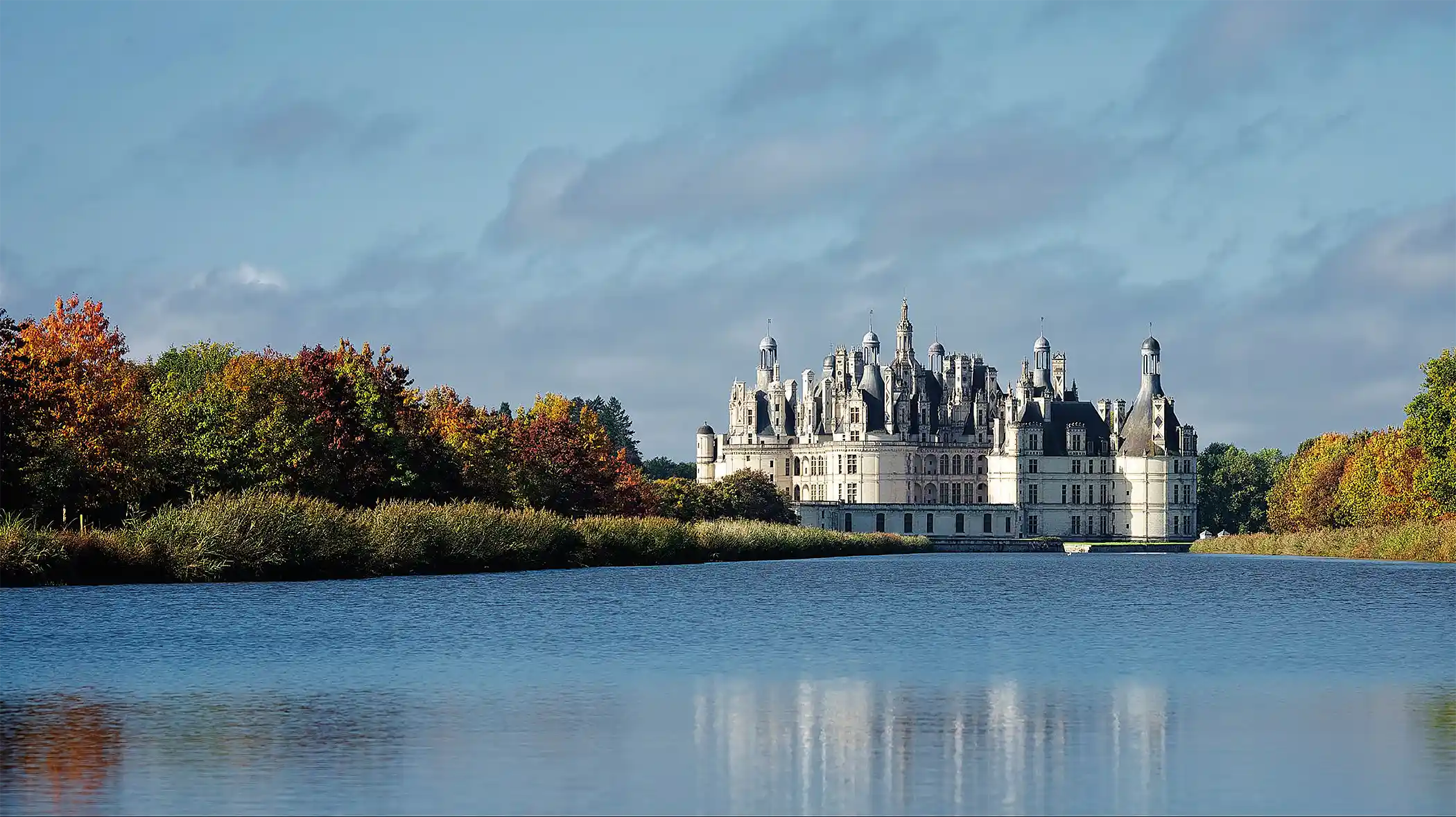 Château blanc entouré d'arbres aux couleurs d'automne avec un grand plan d'eau devant.