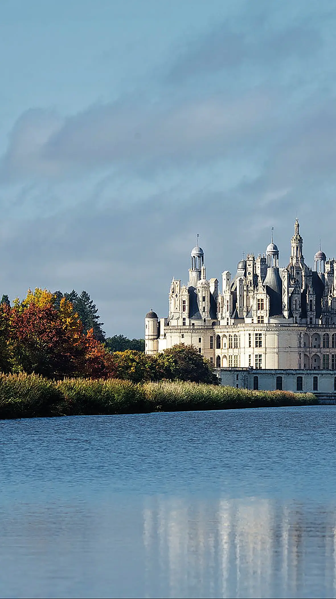 Château blanc avec tours et tourelles vu derrière un étang et des arbres aux feuilles d’automne.
