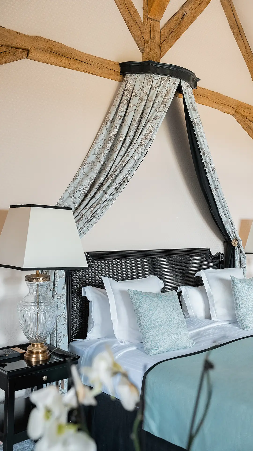 Bett mit schwarzen Kopfteilen, dekorativen Vorhängen, blauen Kissen und Lampe auf schwarzem Nachttisch.