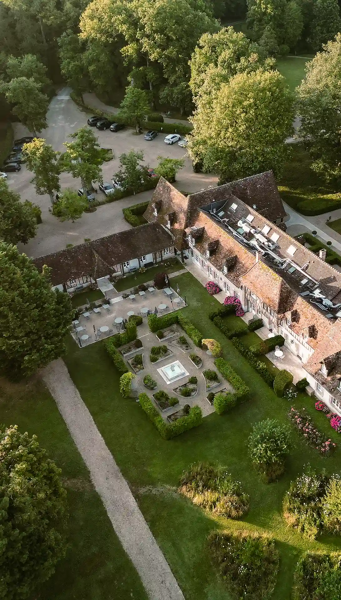 Vue aérienne d'un bâtiment ancien avec patio, jardin structuré, terrasses et voitures garées à l'ombre des arbres.