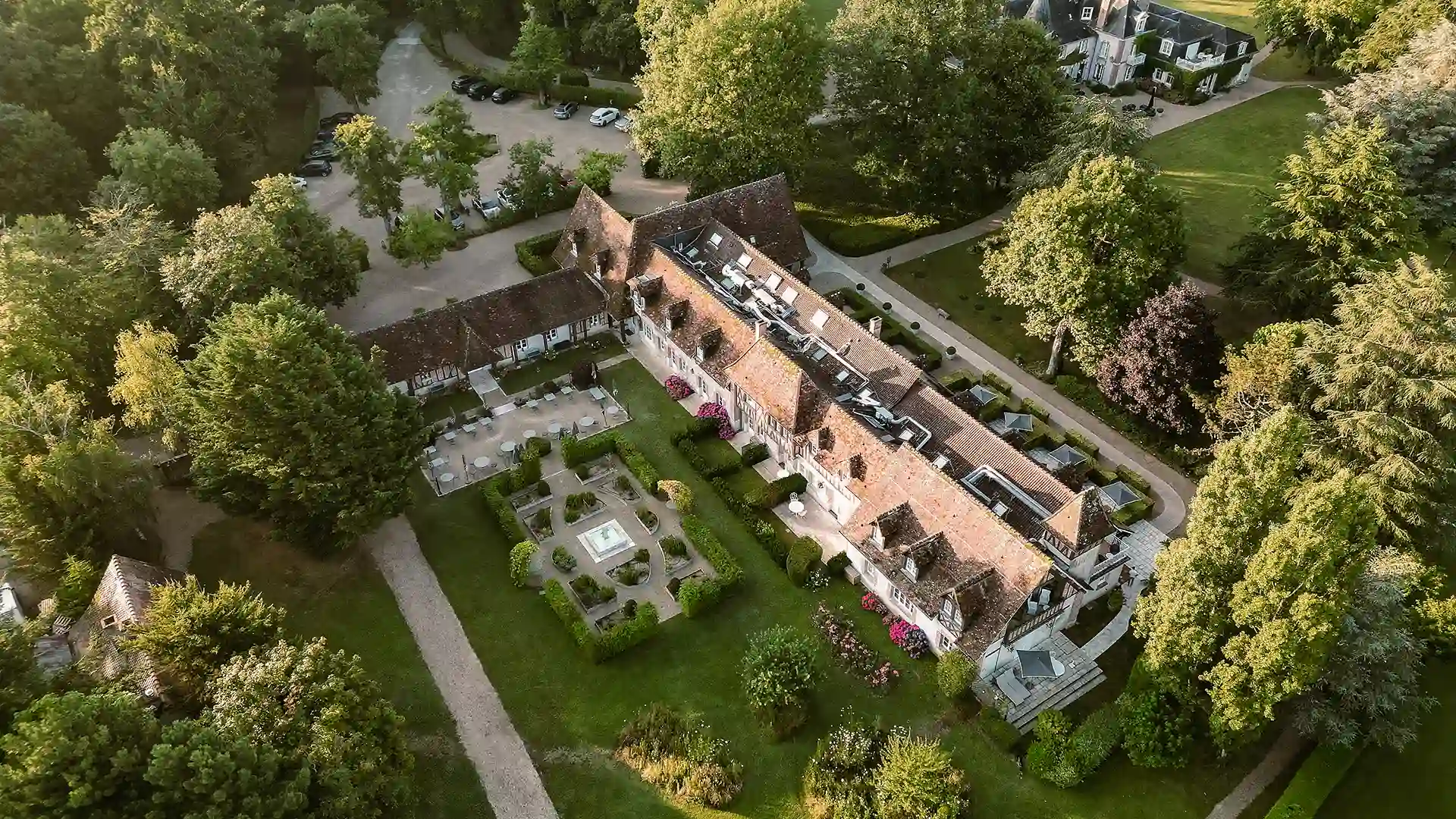 Vue aérienne d'un grand manoir avec un toit en tuiles, entouré de jardins et d'arbres verts.
