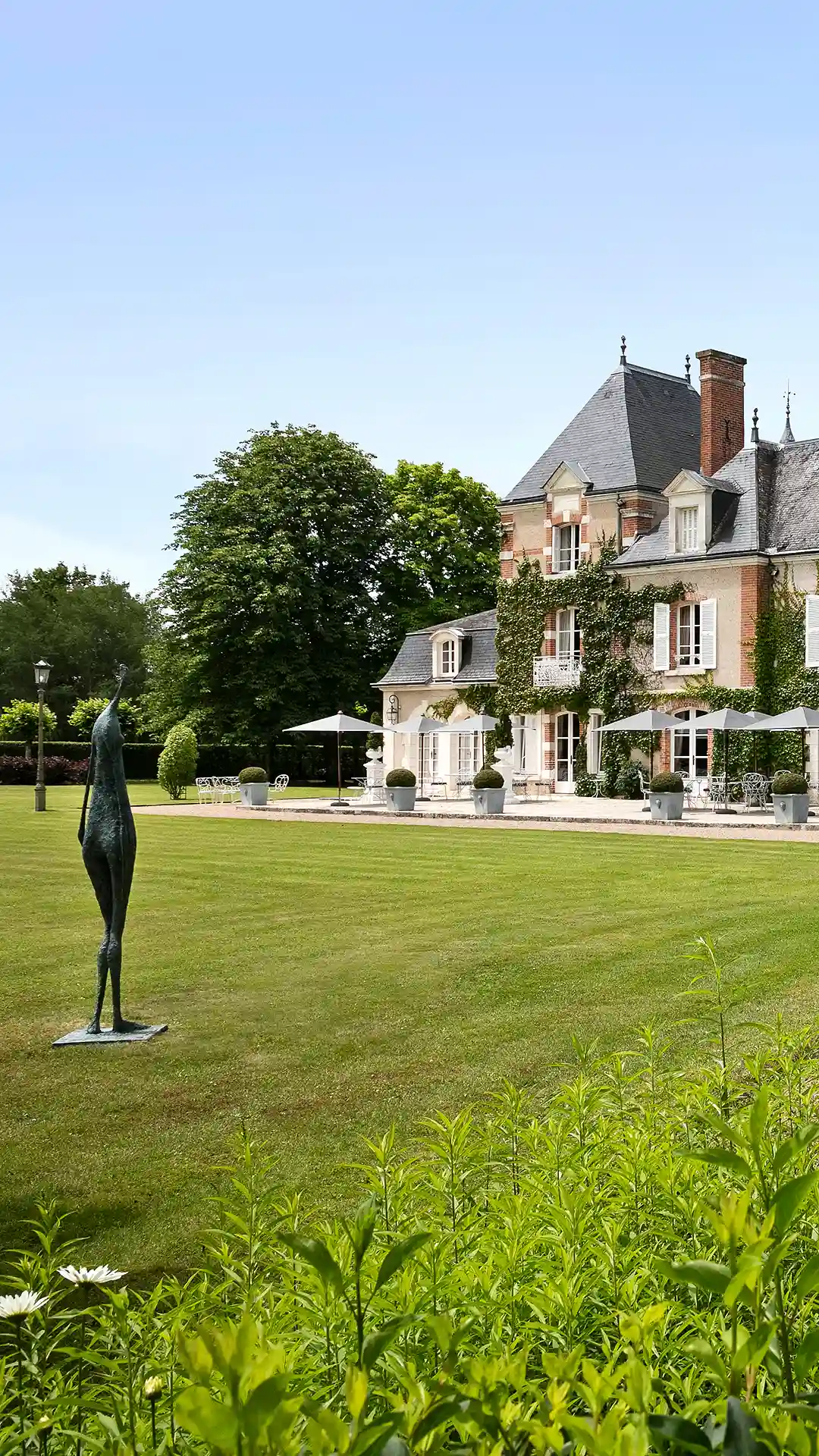 Stone mansion with ivy, manicured lawn and slender statue in the foreground.