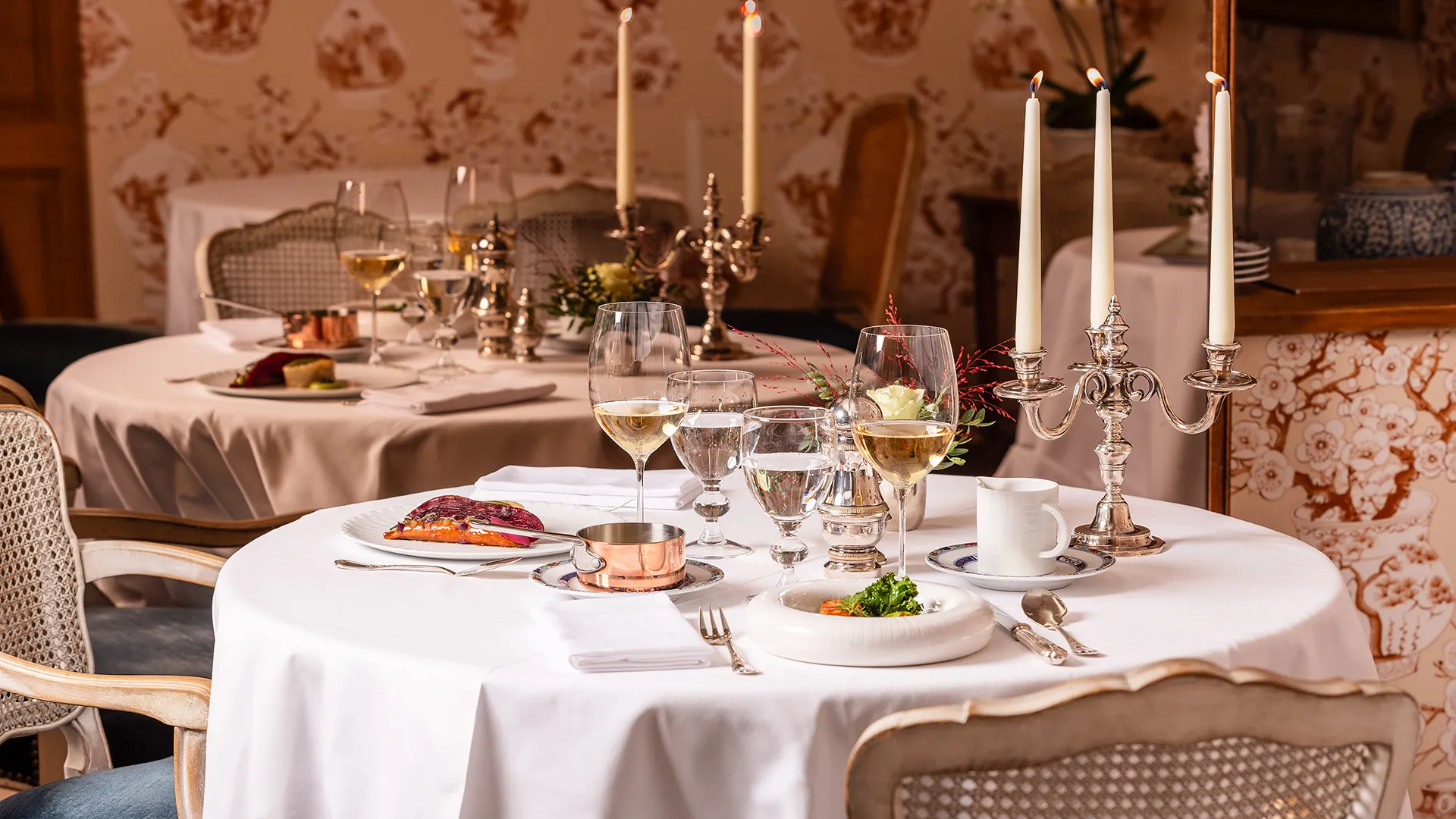 Elegantly set table with white tablecloths, wine glasses, gourmet platter, and three-candle candlestick.