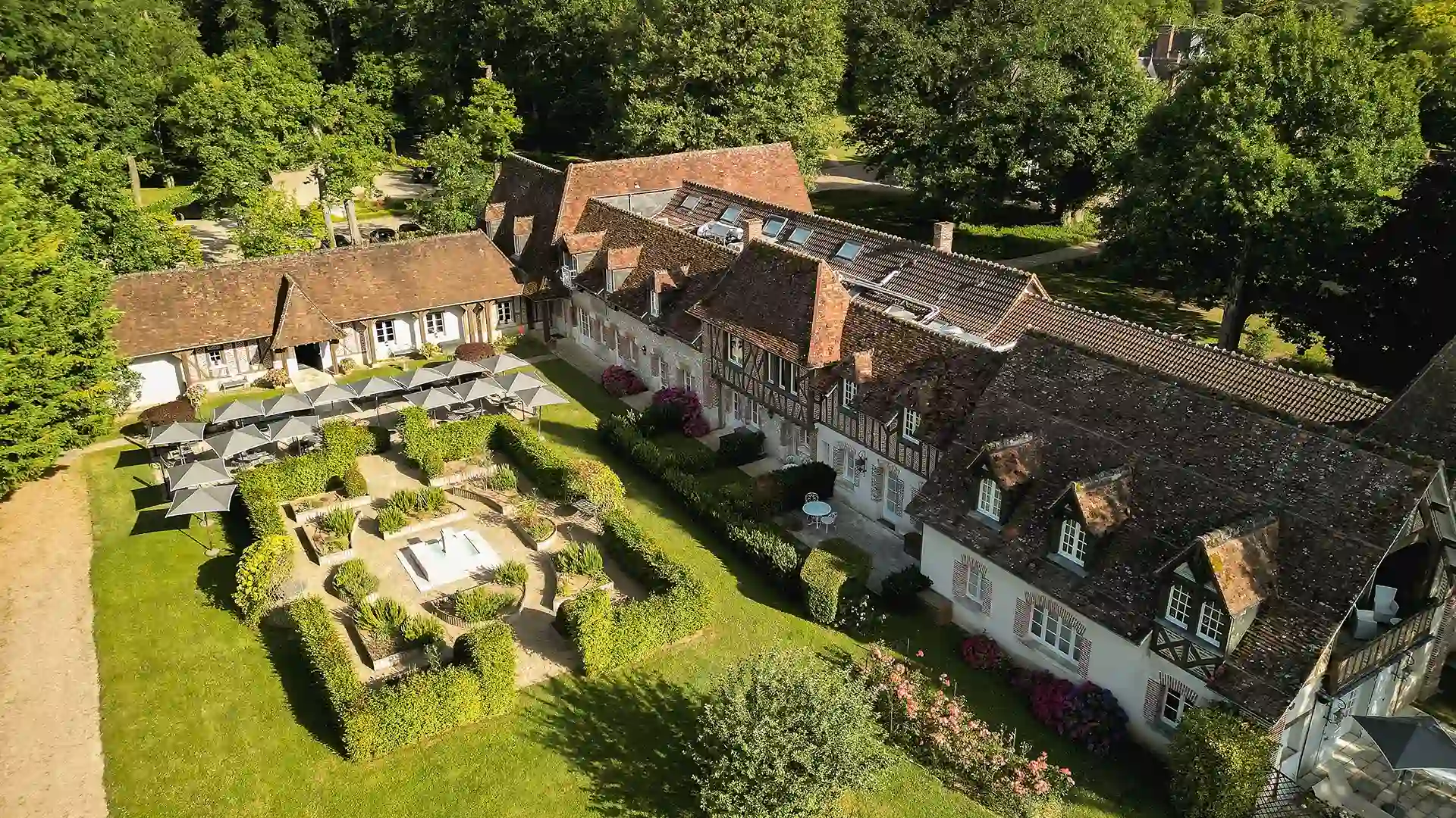 Luftaufnahme eines großen traditionellen Steinhauses mit gepflegtem Garten und grauen Sonnenschirmen.