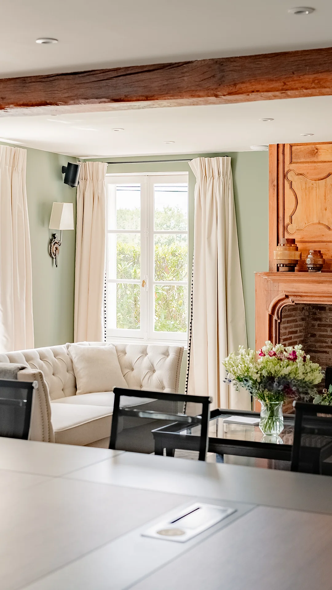 Wohnzimmer mit beigem Sofa, Holzkamin, Fenstern mit Lichtvorhängen und einem Blumenstrauß auf dem Tisch.