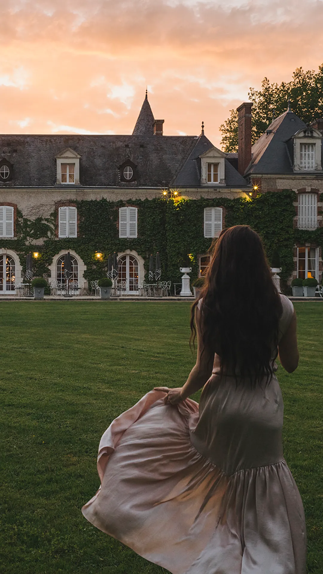 Woman in dress holding her skirt in front of a big old house with green lawn at sunset.