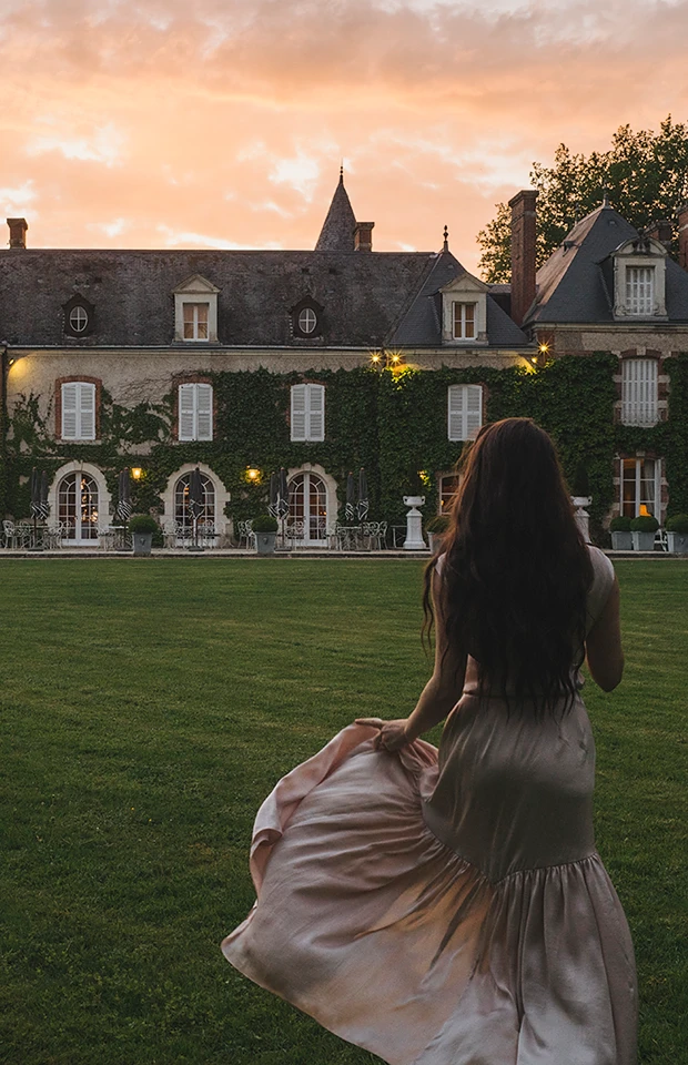 Une femme en robe longue marche sur la pelouse vers un grand manoir couvert de lierre au coucher du soleil.