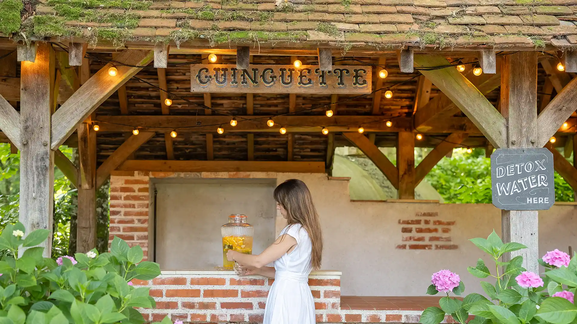 Les Hauts de Loire — Hotel, restaurante y spa — 5 estrellas: mujer con un vestido blanco que sirve agua desintoxicante bajo un refugio de madera iluminado por guirnaldas.