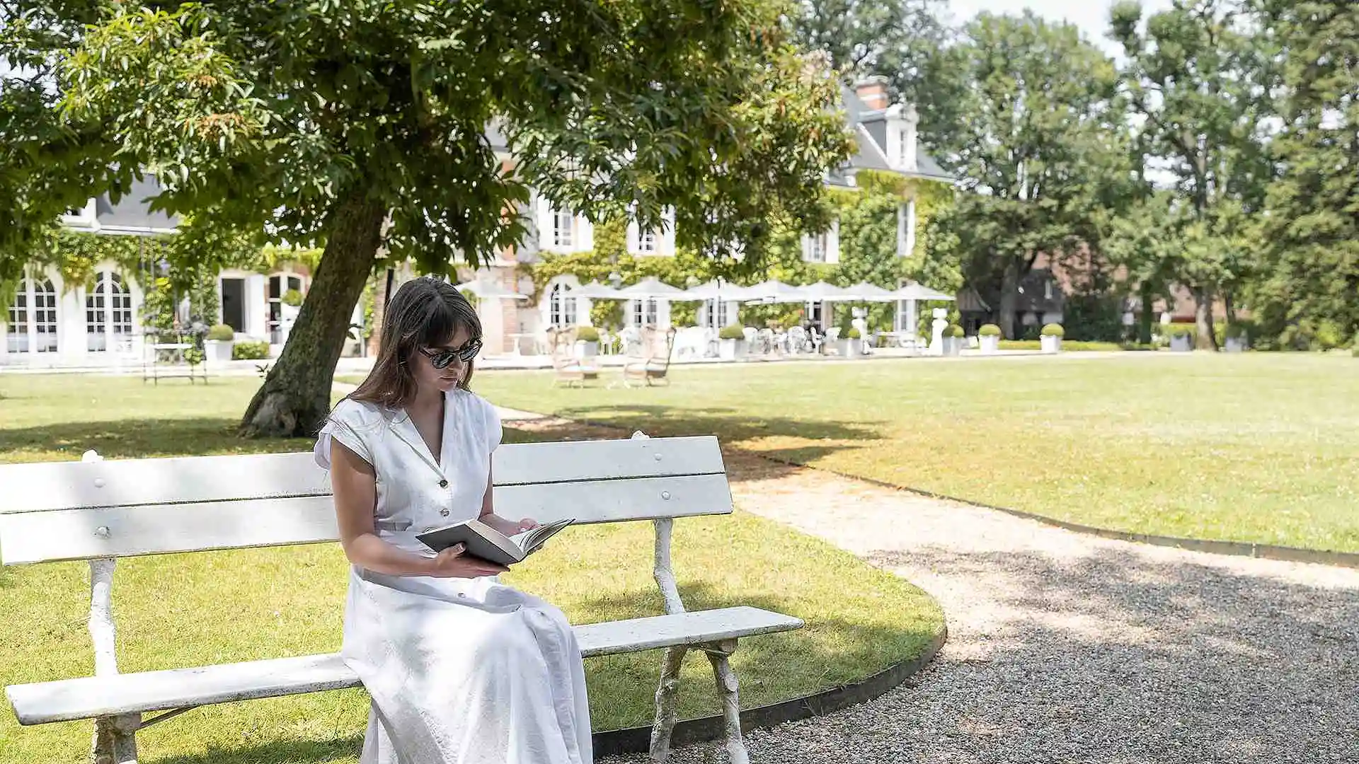 Les Hauts de Loire – Hôtel, Restaurant & Spa – 5 étoiles : femme en robe blanche lisant sur un banc blanc dans un parc ensoleillé.