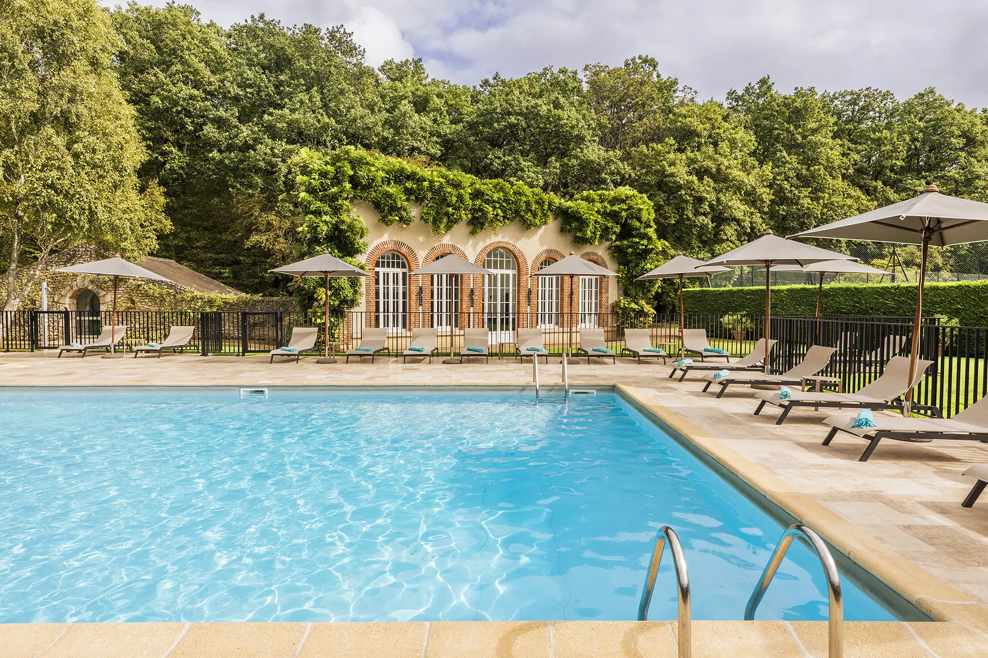 Outdoor pool with sun loungers and umbrellas in front of a building decorated with green plants.