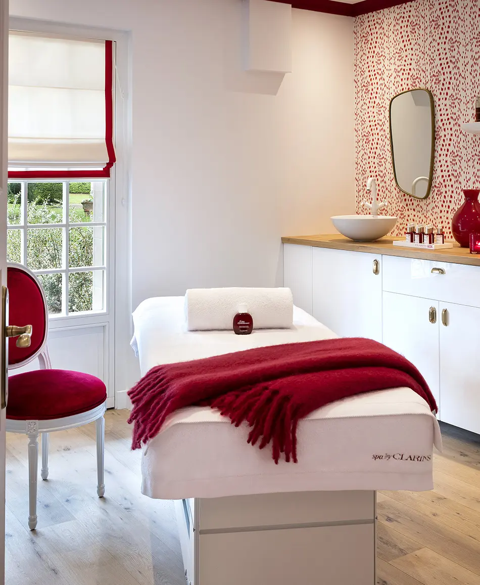 Behandlungsraum mit weißem Tisch, roter Decke, gerolltem Handtuch und rotem Stuhl am Fenster.