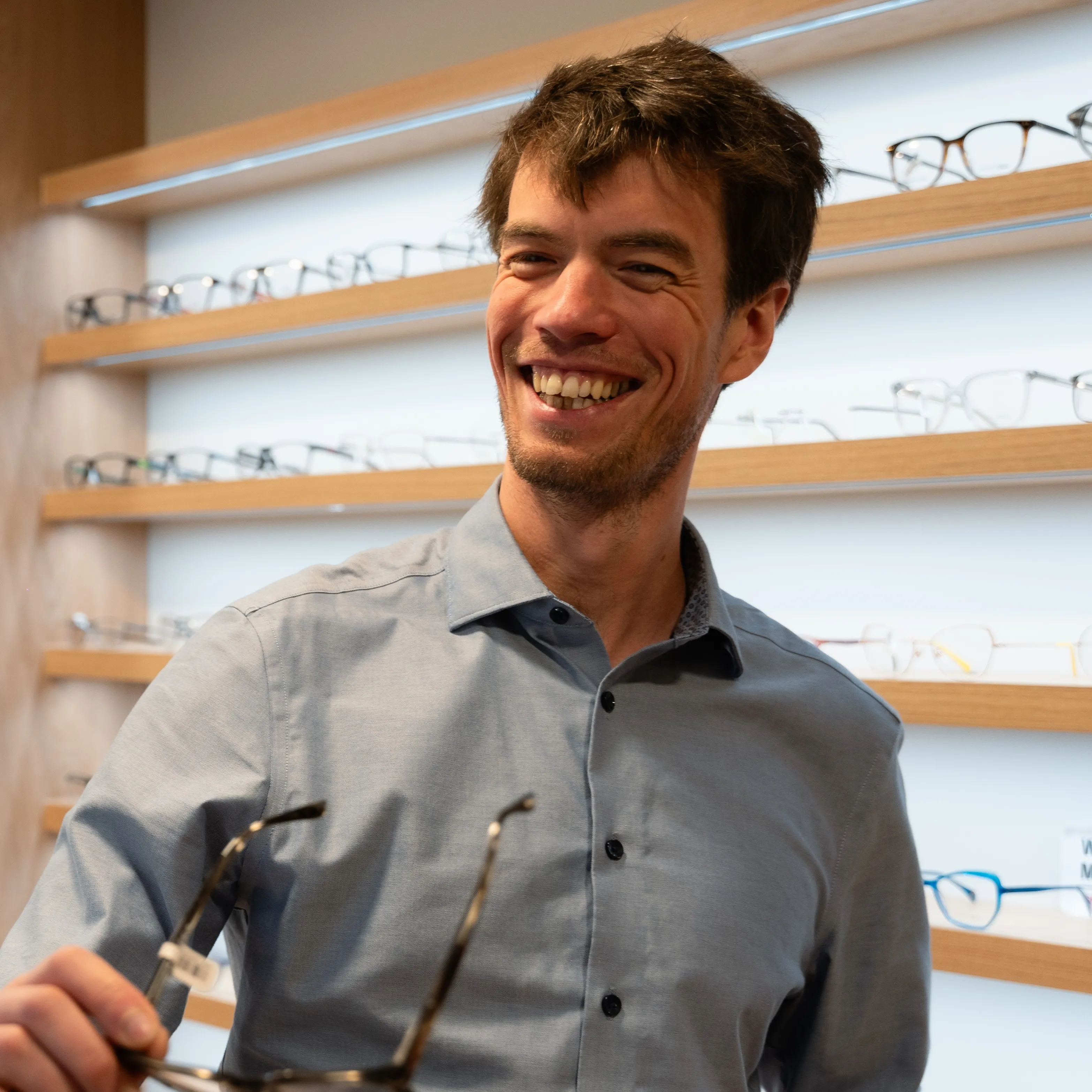 Smiling man holding eyeglasses in an optical store with shelves of glasses behind him.