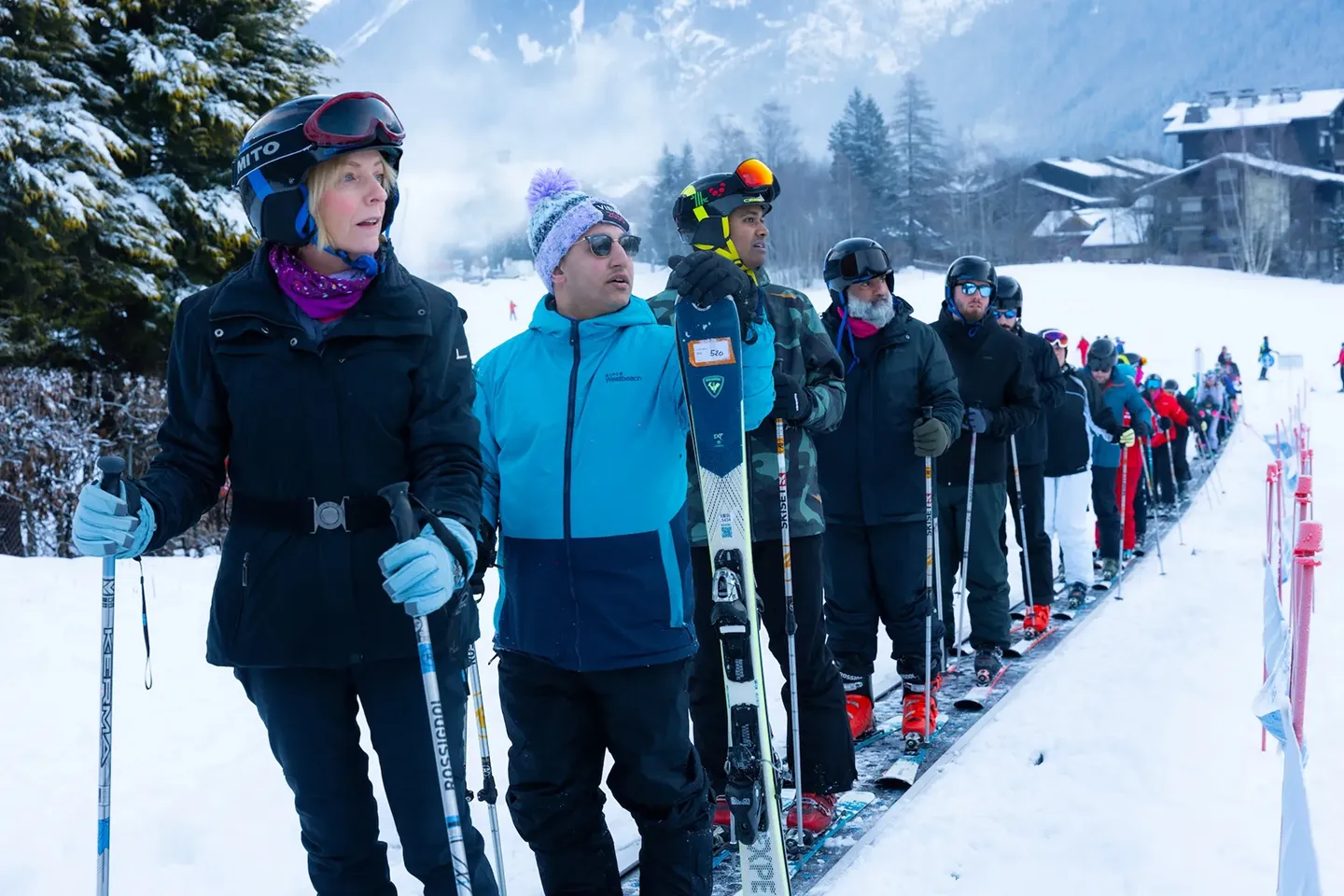 A diverse group of skiers standing in line on a snowy slope, holding ski poles and skis, wearing winter gear and helmets.