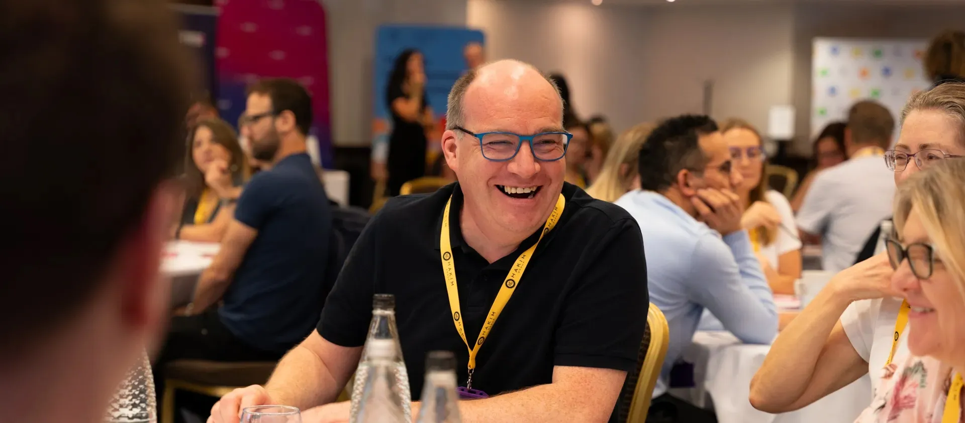 Smiling bald man wearing blue glasses and a yellow lanyard sitting at a table in a lively conference room.