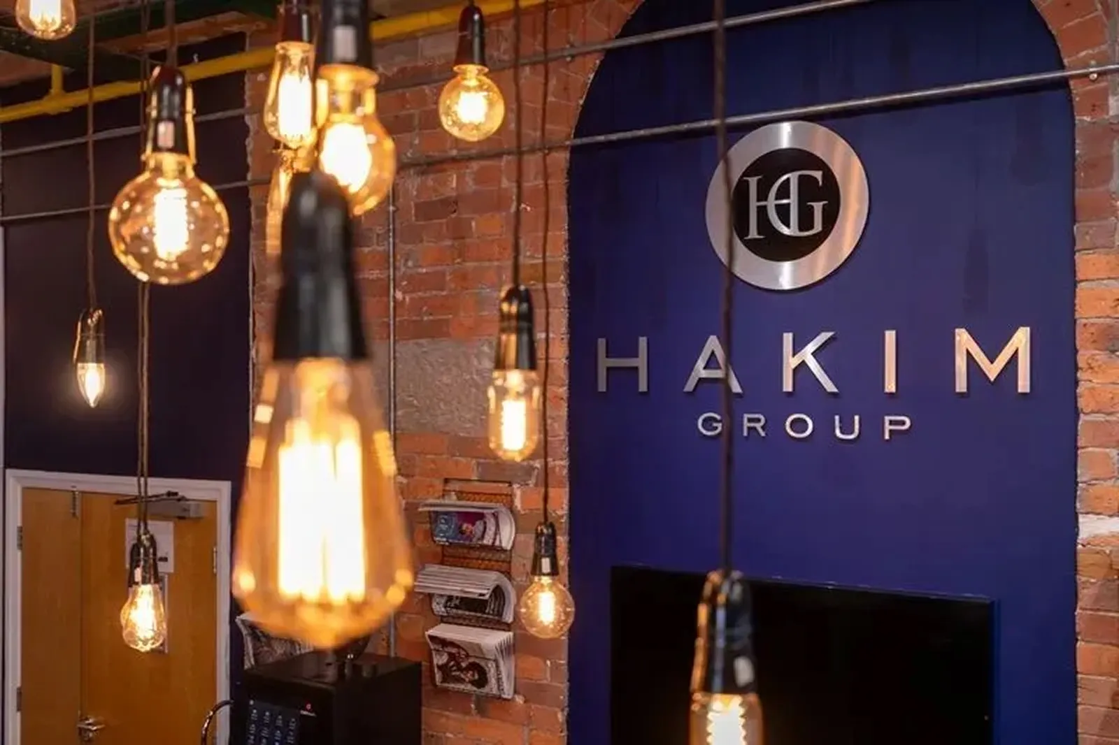 Modern office interior with hanging vintage-style bulbs and a blue wall featuring the Hakim Group logo in silver letters.