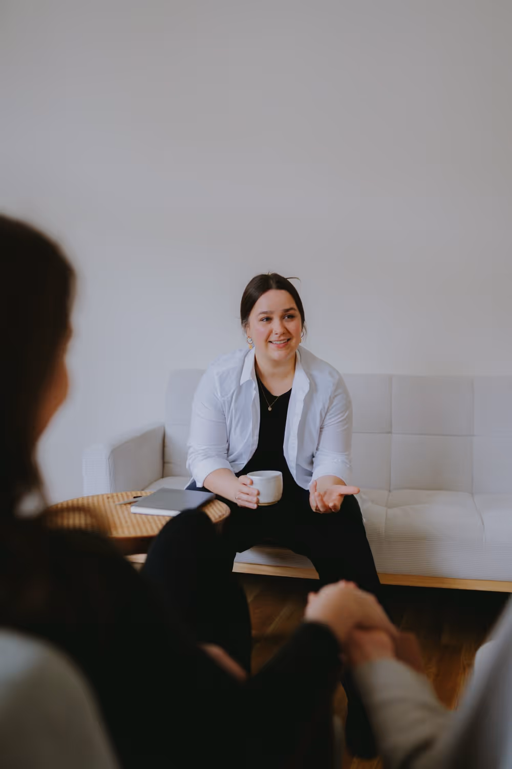 Woman in white shirt holding a cup, sitting on couch and talking to two people holding hands.