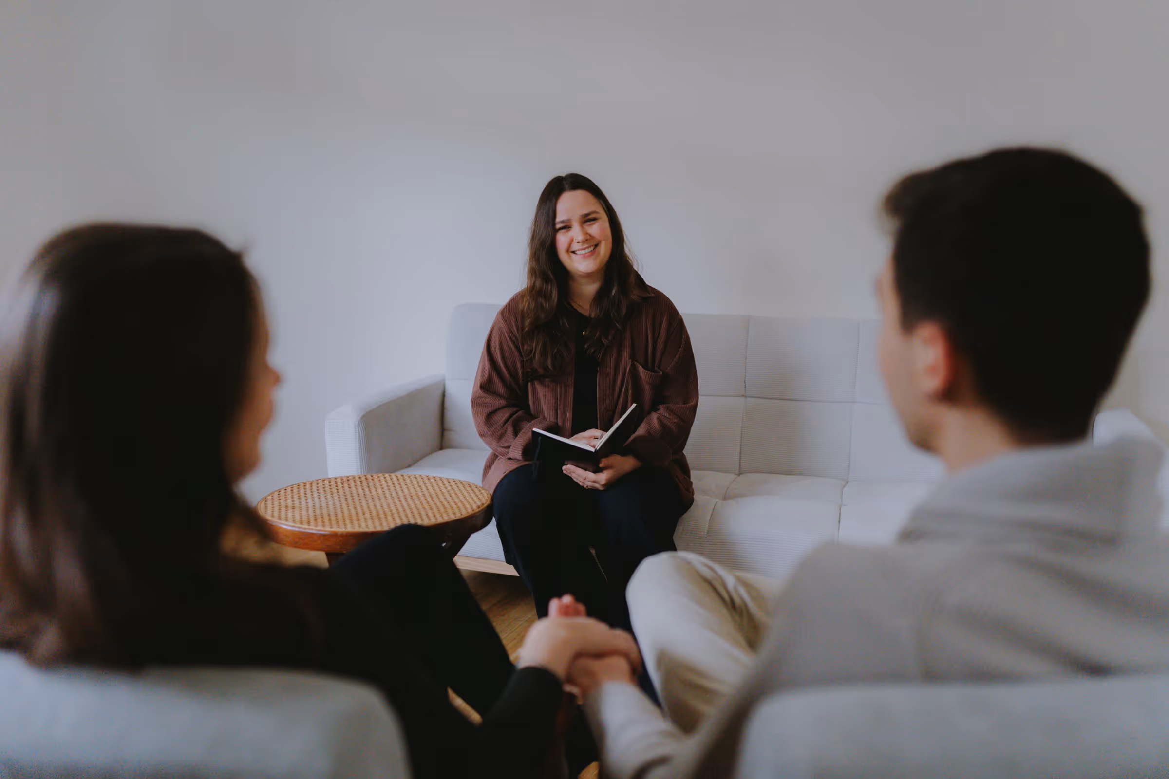 Smiling woman holding a notebook sits on a white couch opposite a couple holding hands.