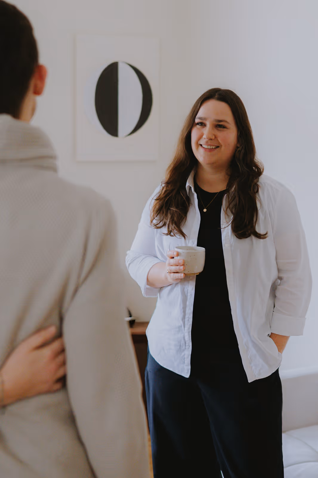 Smiling woman holding a cup talks with a person wearing a beige sweater in a bright room.