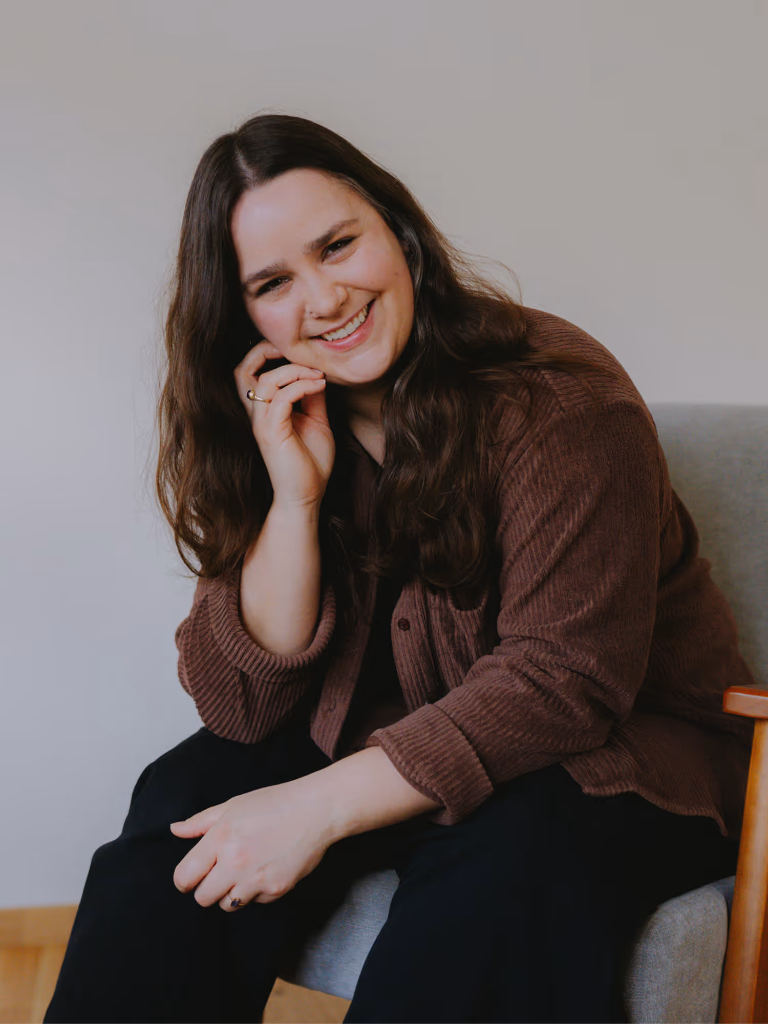 Smiling woman with long dark hair in a brown jacket sitting on a gray chair against a plain wall.