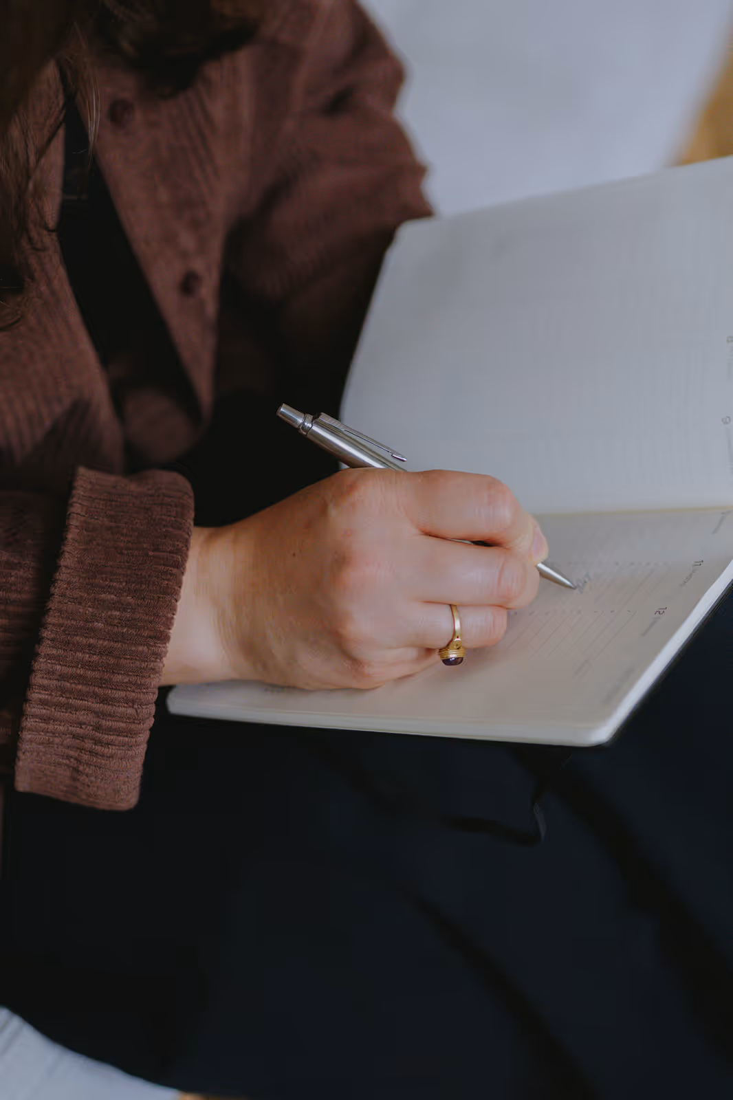 Person in brown sweater writing in a notebook with a silver pen, wearing a gold ring with a purple stone.
