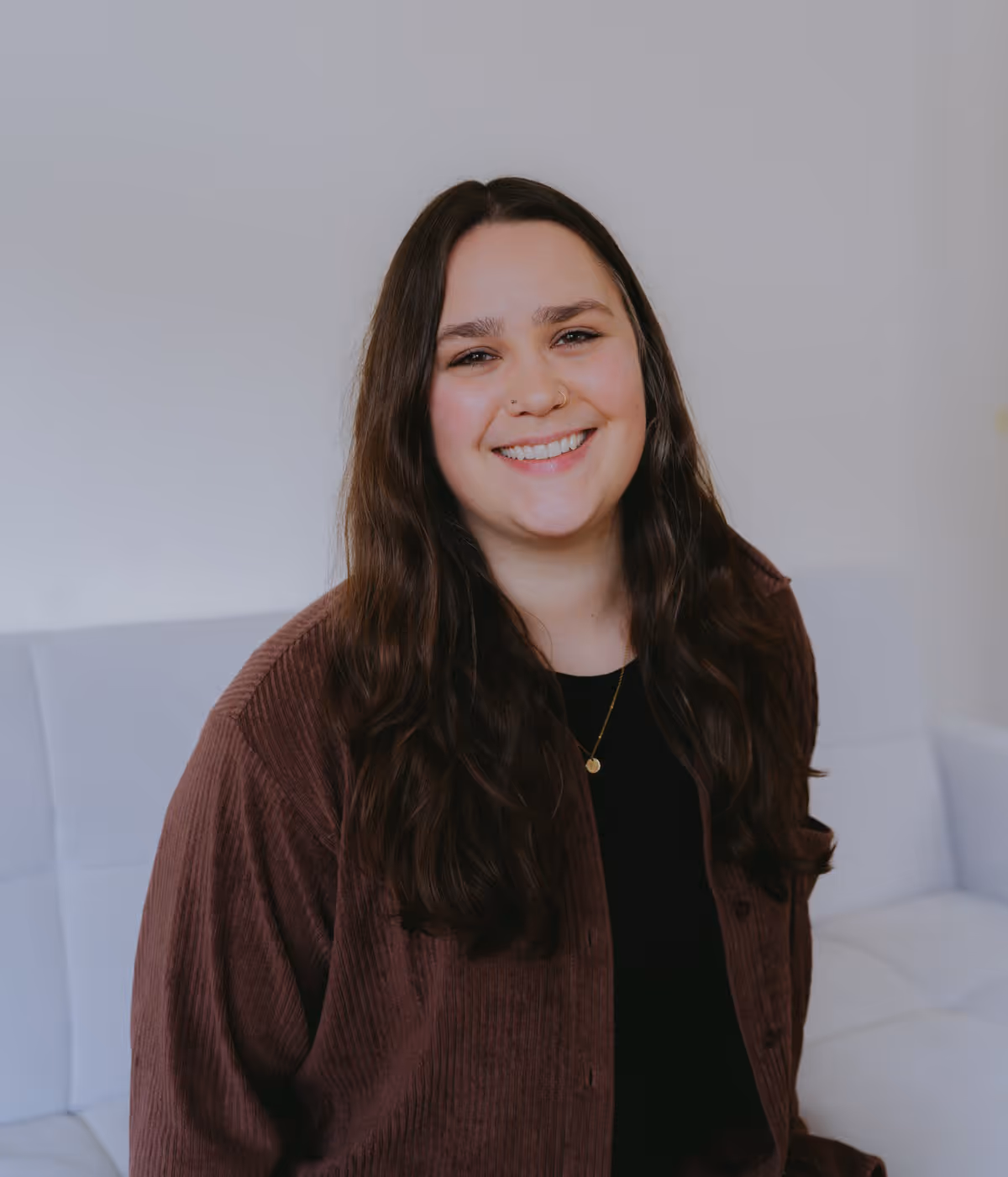 Smiling woman with long dark hair wearing a brown jacket and black top, sitting on a white couch.