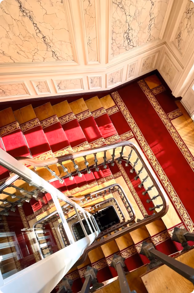 Vue plongeante d'un escalier tournant avec tapis rouge et motif doré dans un bâtiment élégant.