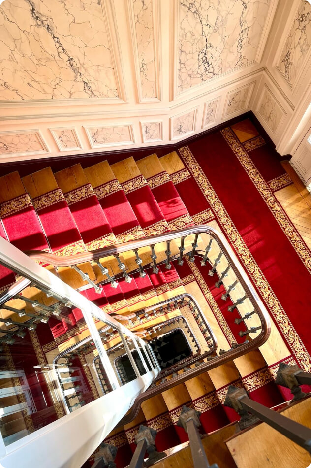 Vue plongeante d'un escalier tournant avec tapis rouge et motif doré dans un bâtiment élégant.