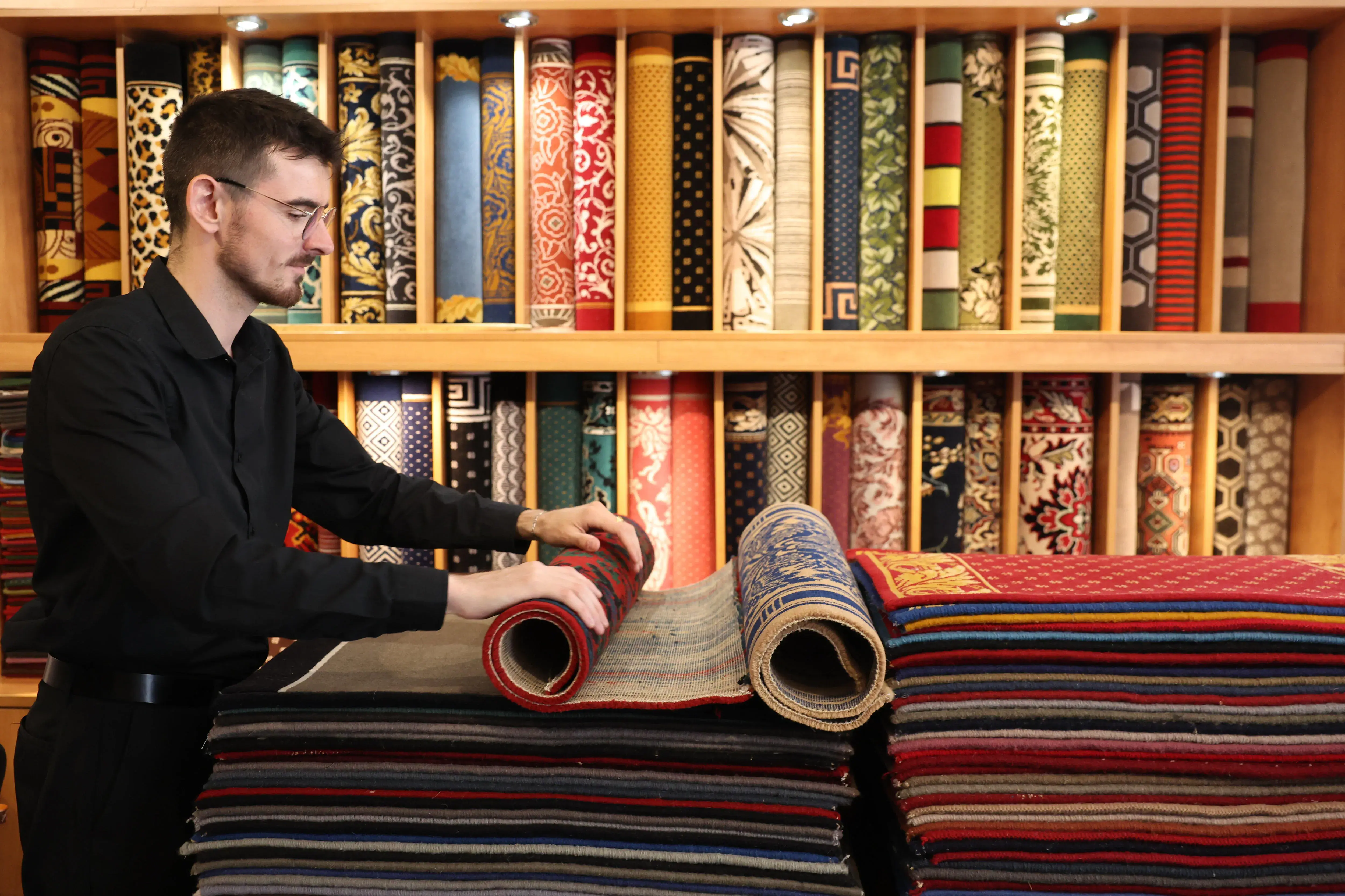 Homme déroulant un tapis coloré dans un magasin avec des rouleaux de tapis décoratifs en arrière-plan.