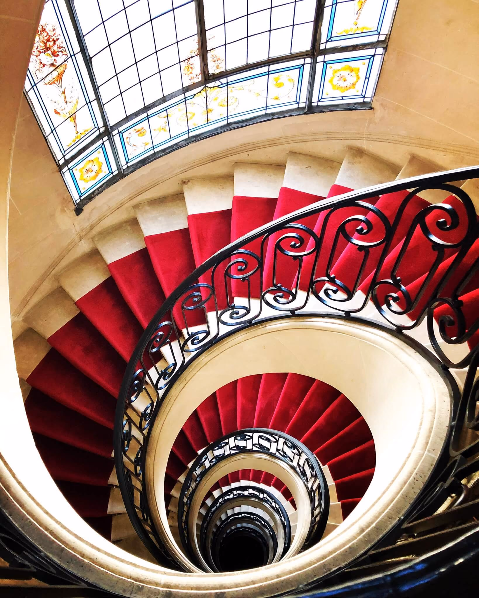 Vue plongeante d'un escalier en colimaçon avec tapis rouge et balustrade en fer noir orné.