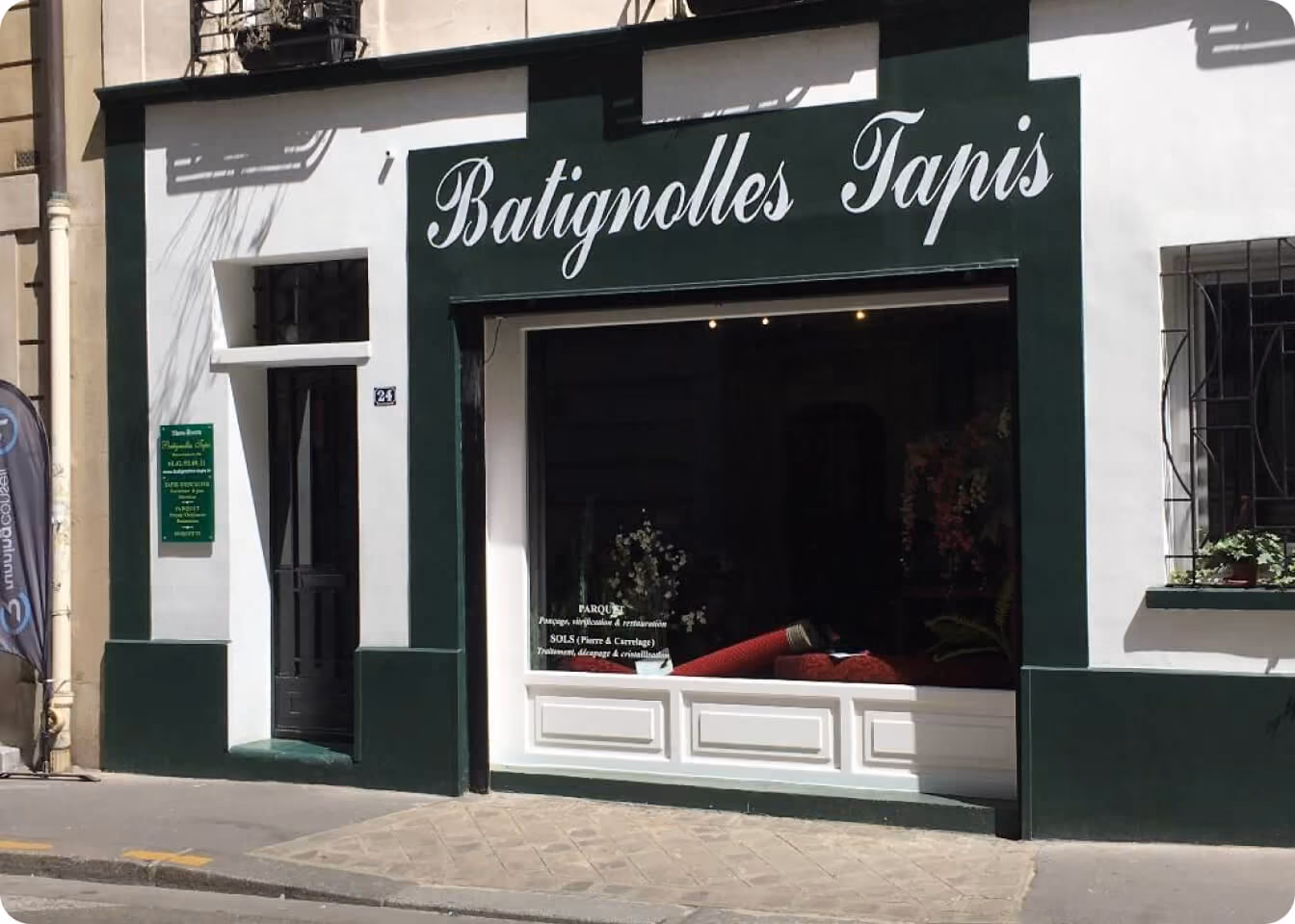 Façade d'un magasin nommé Batignolles Tapis avec une vitrine affichant des tapis rouges et plantes.