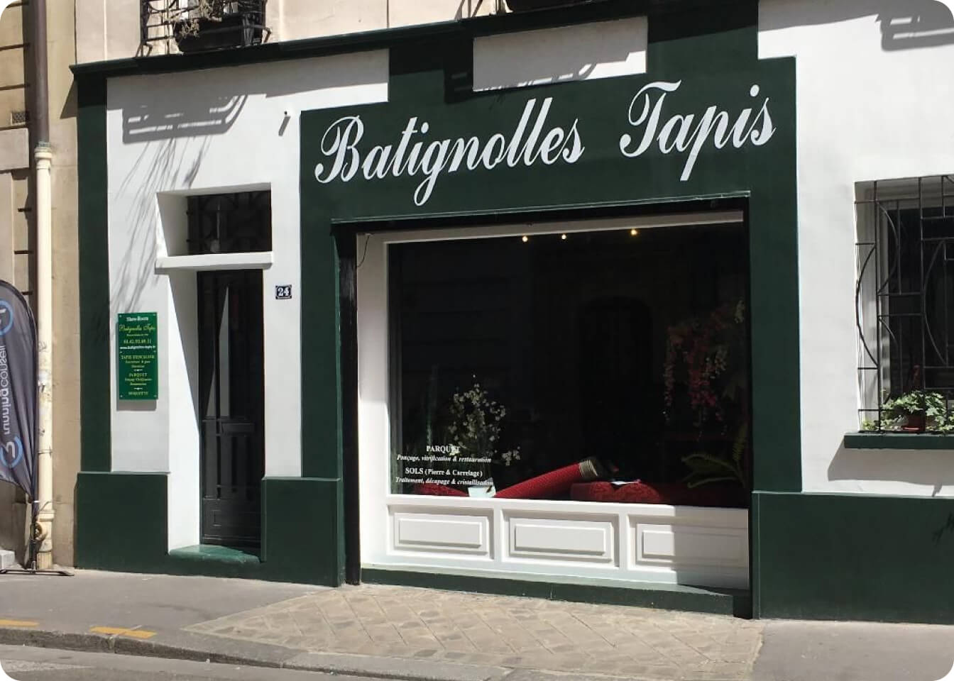Façade d'un magasin nommé Batignolles Tapis avec une vitrine affichant des tapis rouges et plantes.