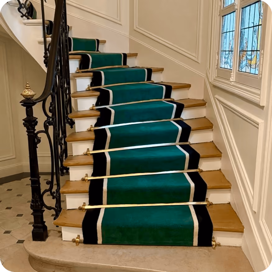 Escalier en bois avec un tapis vert à bordures noires et blanches fixé par des barres métalliques dorées.