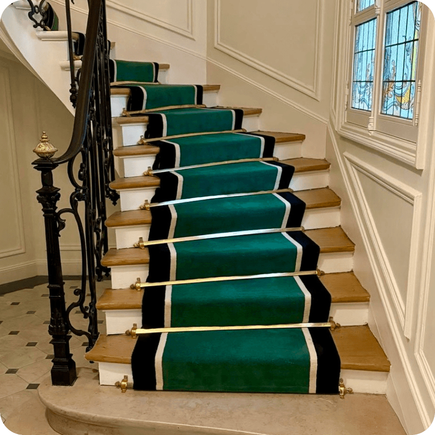 Escalier en bois avec un tapis vert à bordures noires et blanches fixé par des barres métalliques dorées.