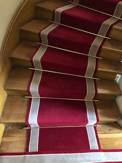 Escalier en bois avec un tapis rouge central bordé de blanc fixé par des barres dorées.